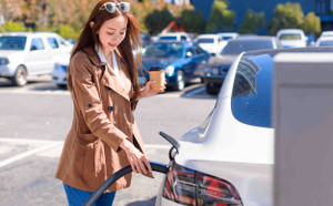 Smiling young woman standing on city parking near electric car, charging automobile battery from small city station. 