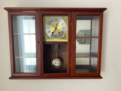 It features a glass-fronted cabinet design with two side compartments. The clock face has a gold-ton...