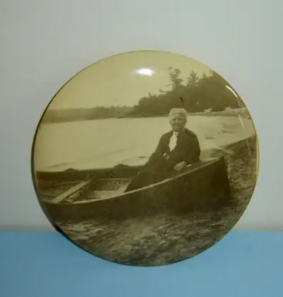 This is a celluloid topped tin 'button' portrait of an elegantly dressed lady sitting in the front o...