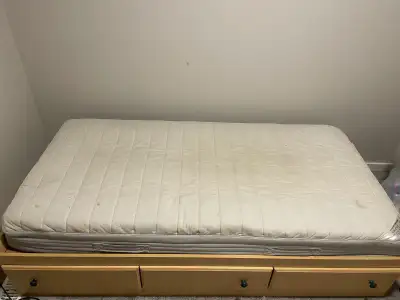 This is a used mattress and a wooden bed frame with three drawers. The mattress is off-white with vi...