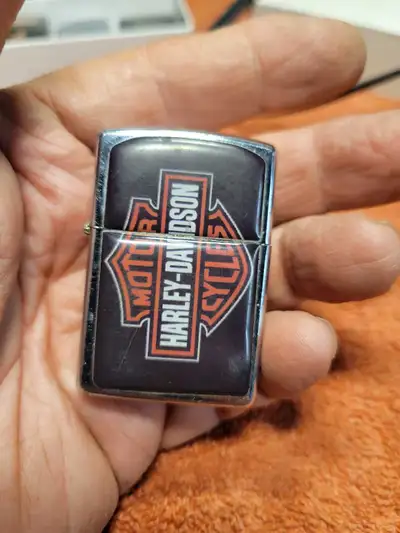 Vintage chrome flip-top lighter with a black and orange Harley-Davidson logo. This metal lighter has...