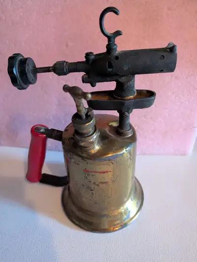 This is an antique blowtorch. Brass body, a red handle, and some signs of wear. It's a piece of hist...