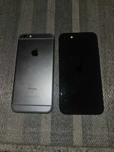 These two Apple iPhones, an iPhone 6s and an iPhone 7, are in poor condition with heavily cracked sc...