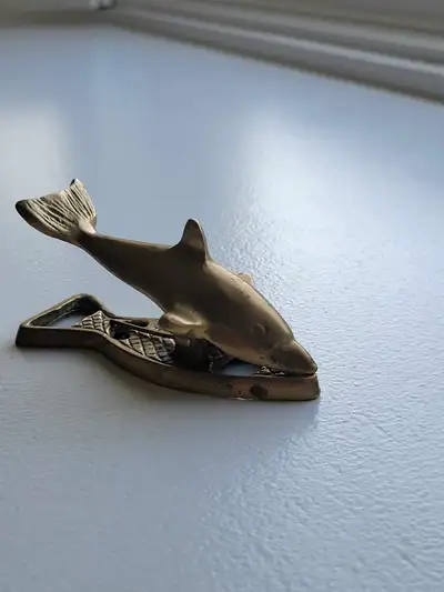 The item is a vintage brass dolphin-shaped bottle opener and paper clip. It functions as a bottle op...