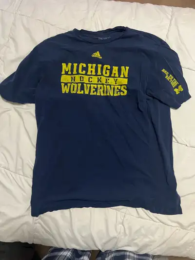 This is a pre-owned Adidas Michigan Hockey Wolverines T-shirt in navy blue and yellow, featuring a c...