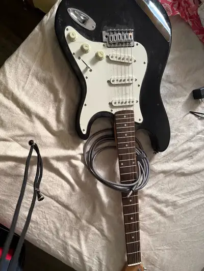 This Squier Stratocaster electric guitar features a classic black finish and a white pickguard, offe...