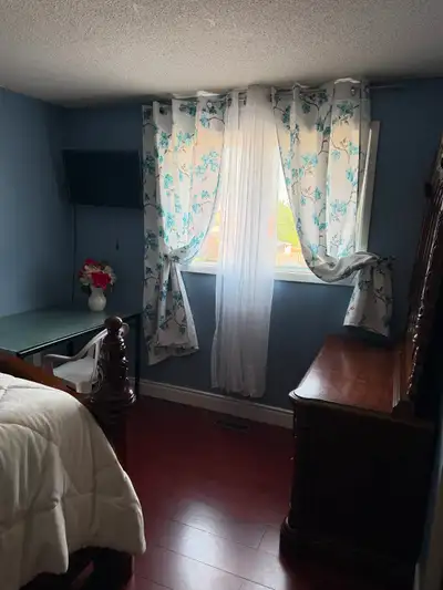 This cozy and well-lit furnished room is available for rent, featuring a comfortable bed, a wooden d...