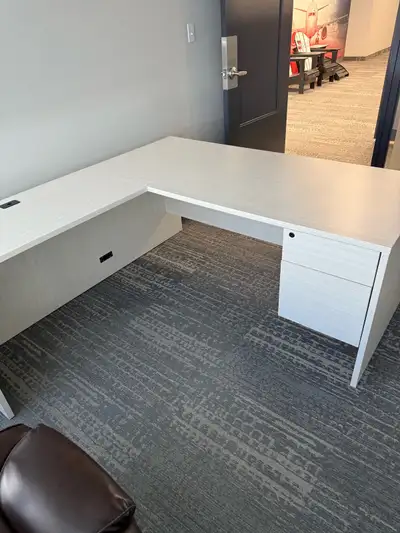 Spacious L-shaped office desk in a light wood grain finish. Desk has a built-in drawer unit. Left re...